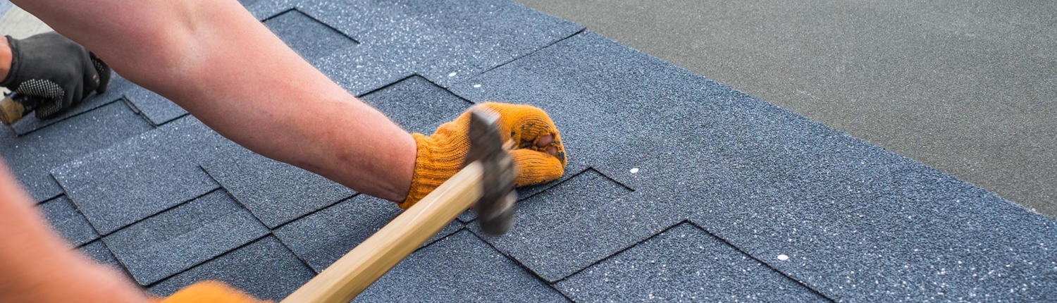 Side view of hand hammering down a nail into the shingles of a roof