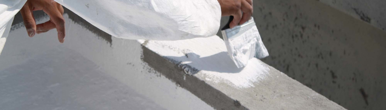 Side view of a hand holding a large paint brush and painting a concrete roof with white paint