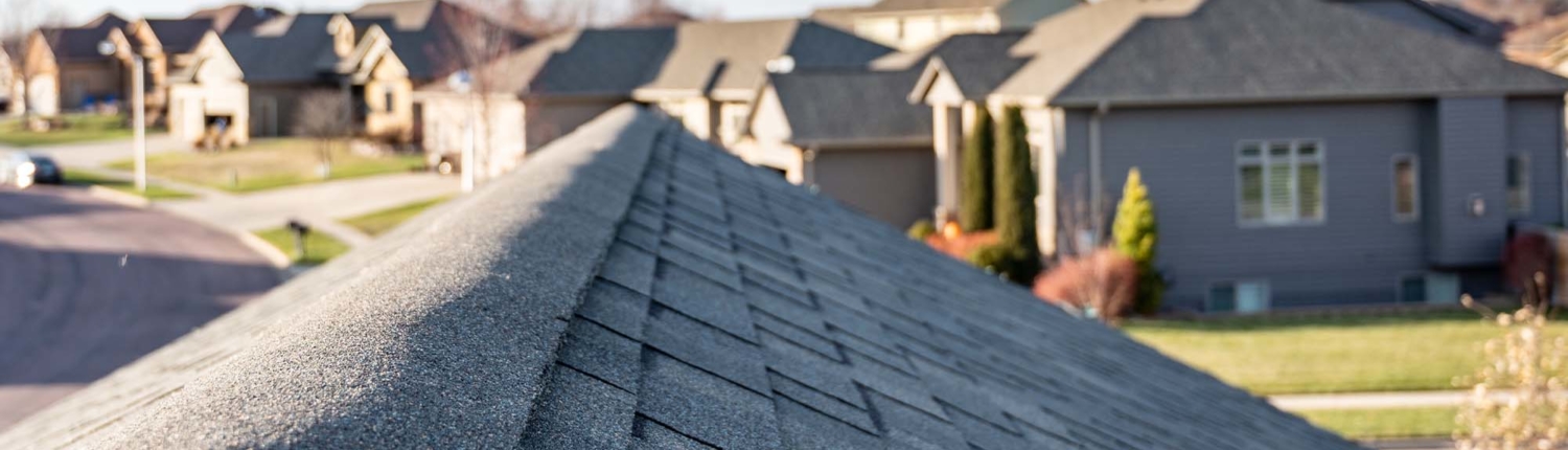 Close up of dark gray shingles on a roof with a neighborhood in the background